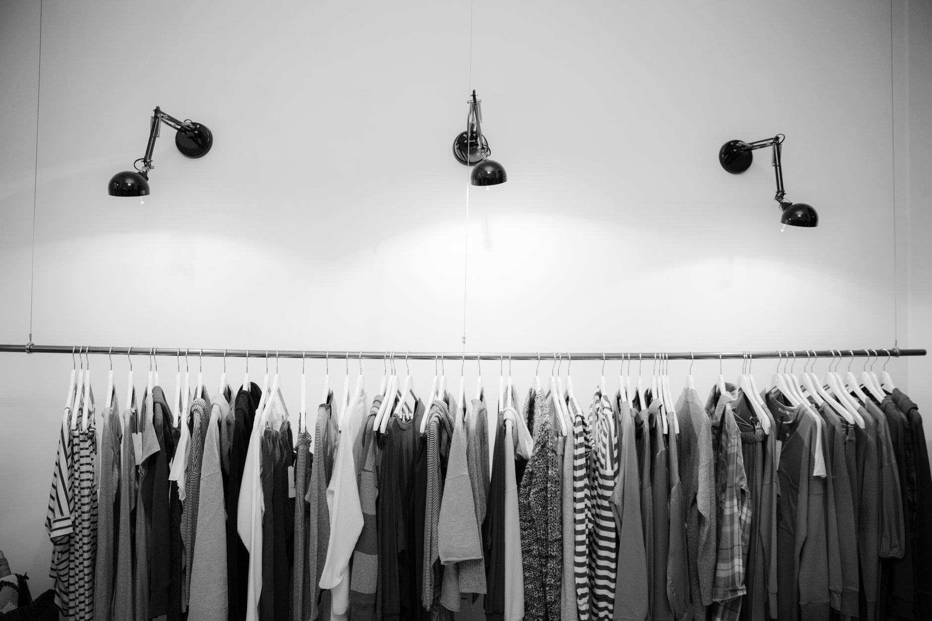 Photo en noir et blanc : vêtements sur cintres le long d’une tringle, sous trois lampes murales articulées, devant un mur clair.